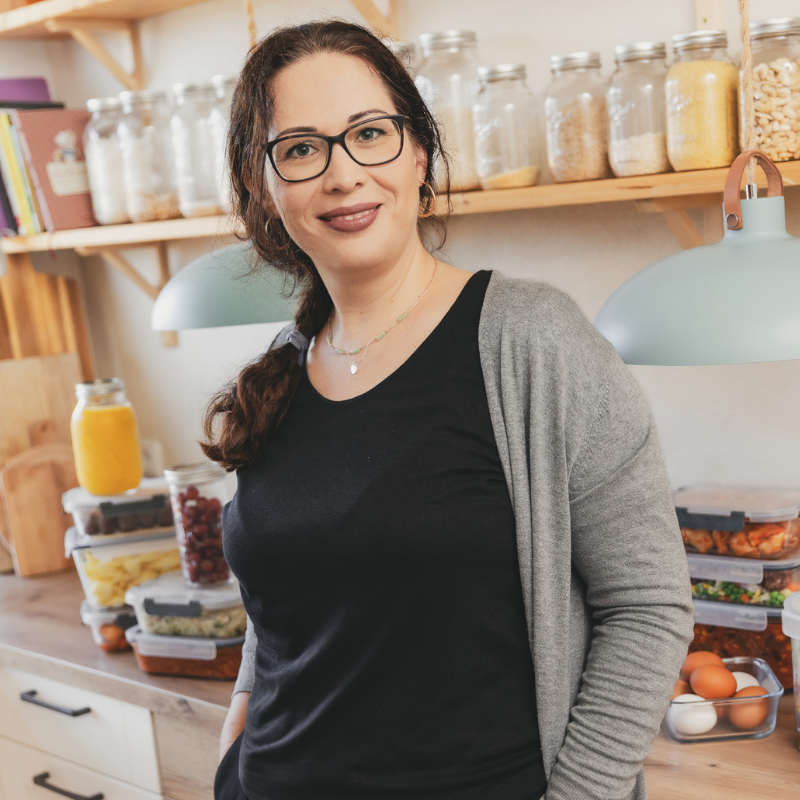 Frau in moderner Küche vor Mealprep-Boxen und Vorratsgläsern, lächelnd in die Kamera.
